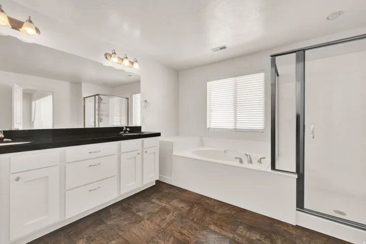 Bathroom featuring a dual vanity with dark countertops, a large soaking tub, a separate glass-enclosed shower, and wood-finish flooring