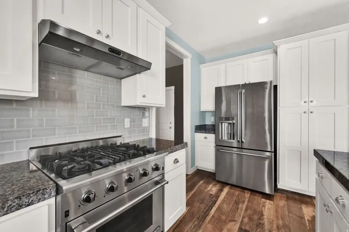 Modern kitchen featuring a stainless steel range with overhead ventilation, light subway tile backsplash, white shaker cabinetry, dark granite countertops, and wood-finish flooring
