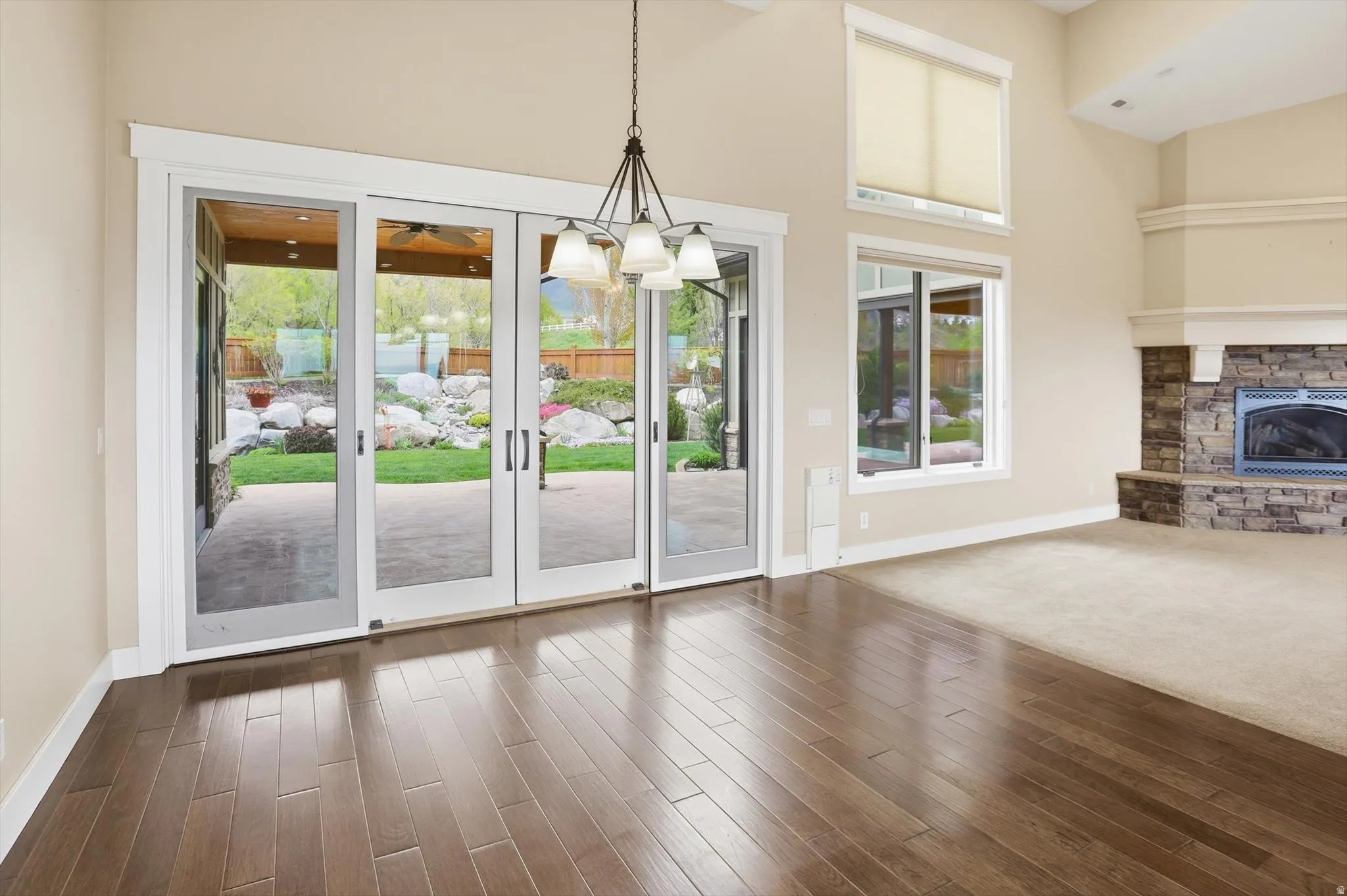 Expansive great room featuring rich wood-finish flooring, a contemporary chandelier, a stone-surround fireplace, and large glass doors opening to a covered patio
