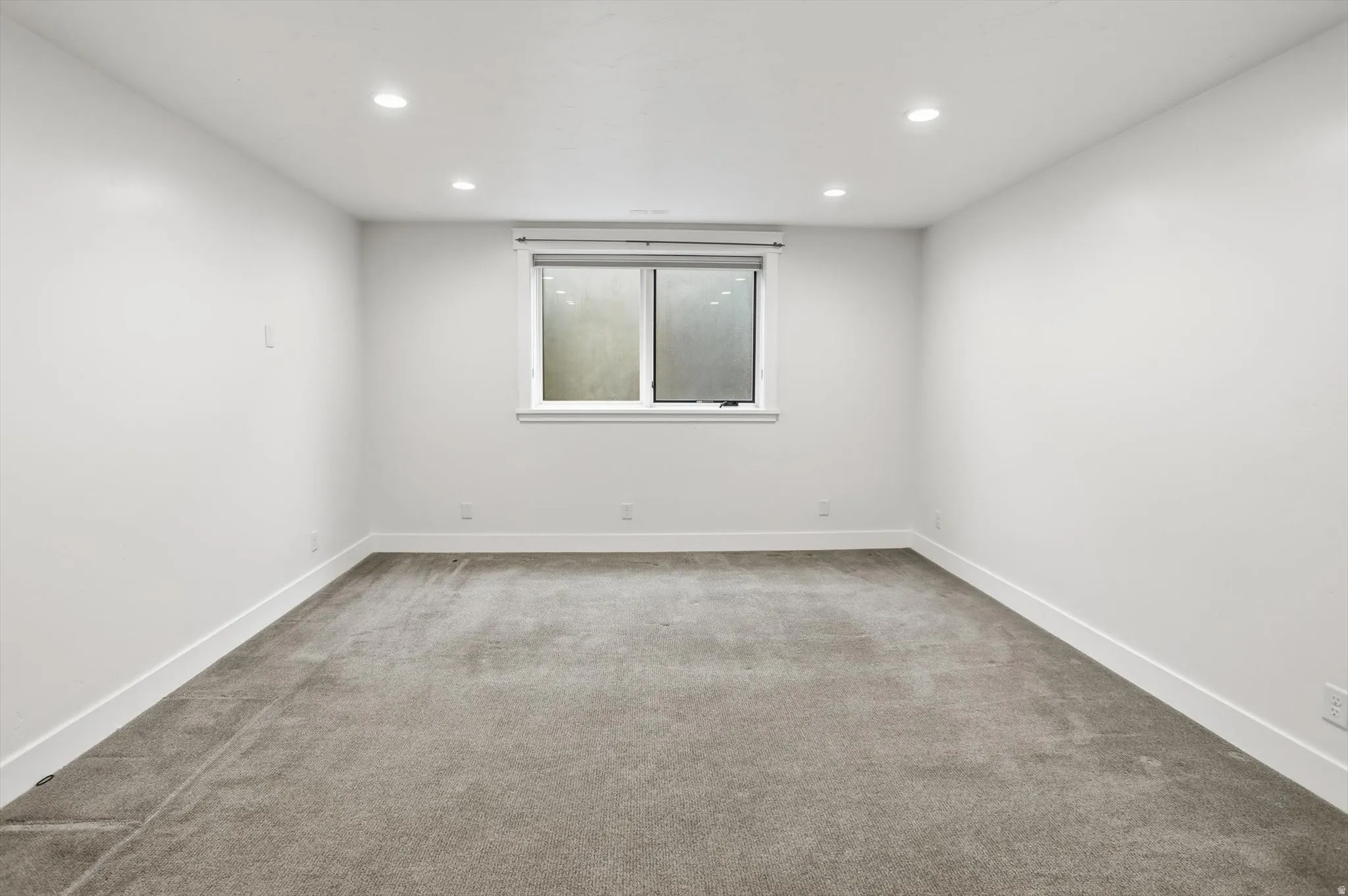 Spacious room featuring neutral grey carpeting, white walls, recessed ceiling lighting, and a window with blinds
