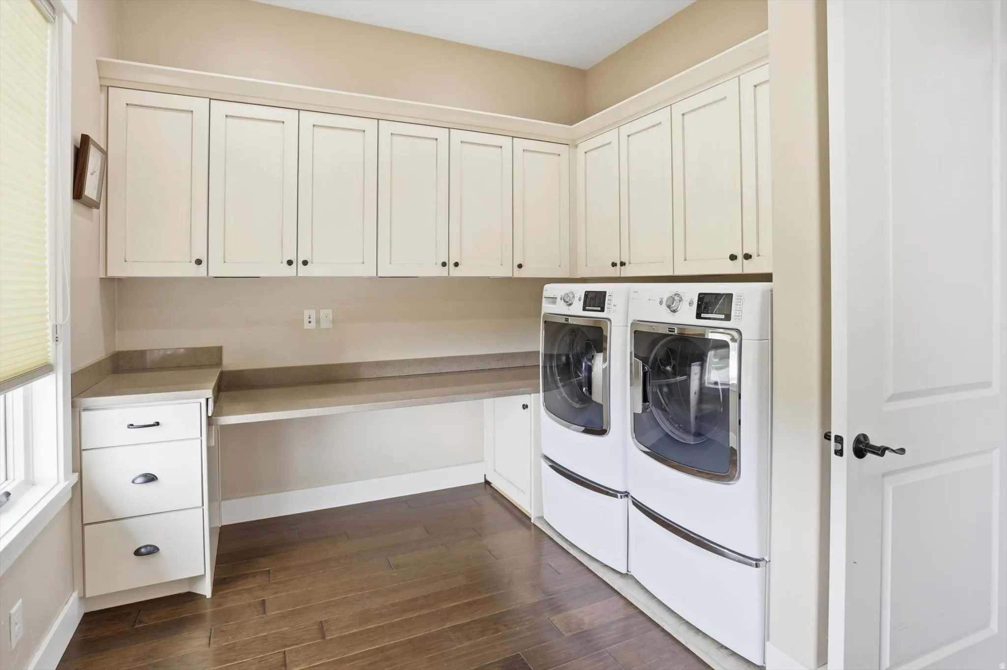 Dedicated laundry space featuring extensive upper cabinetry, stone-finish countertops, built-in lower drawers, wood-finish flooring, and natural light from a window