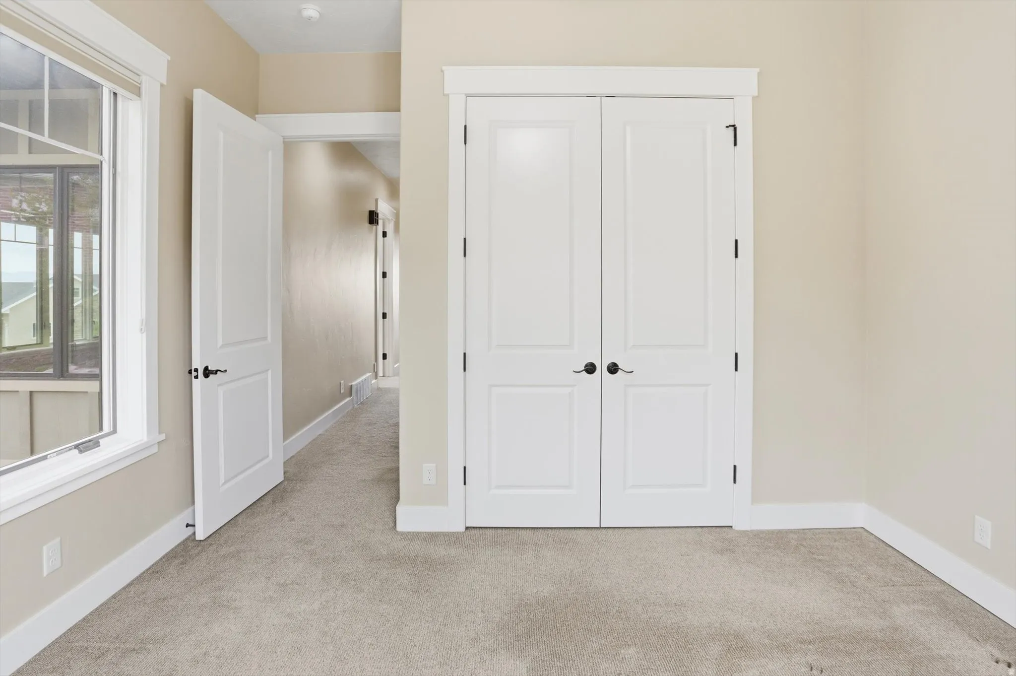 Neutral-toned room featuring a window with white trim, soft grey carpet, and white bi-fold doors with black hardware