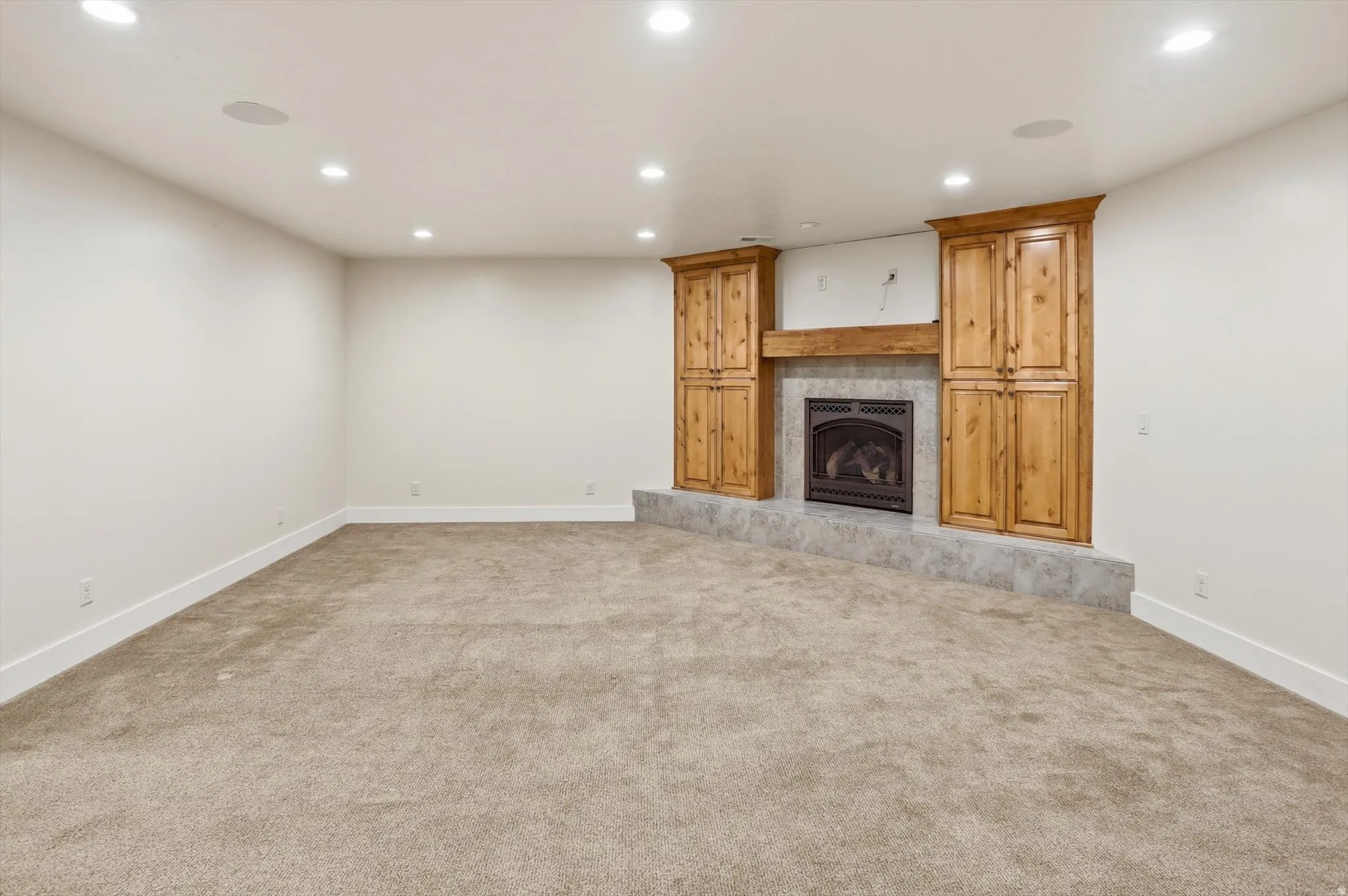 Recessed lighting illuminates a carpeted room featuring a fireplace with a black insert, surrounded by a stone-look hearth and wood-tone cabinetry