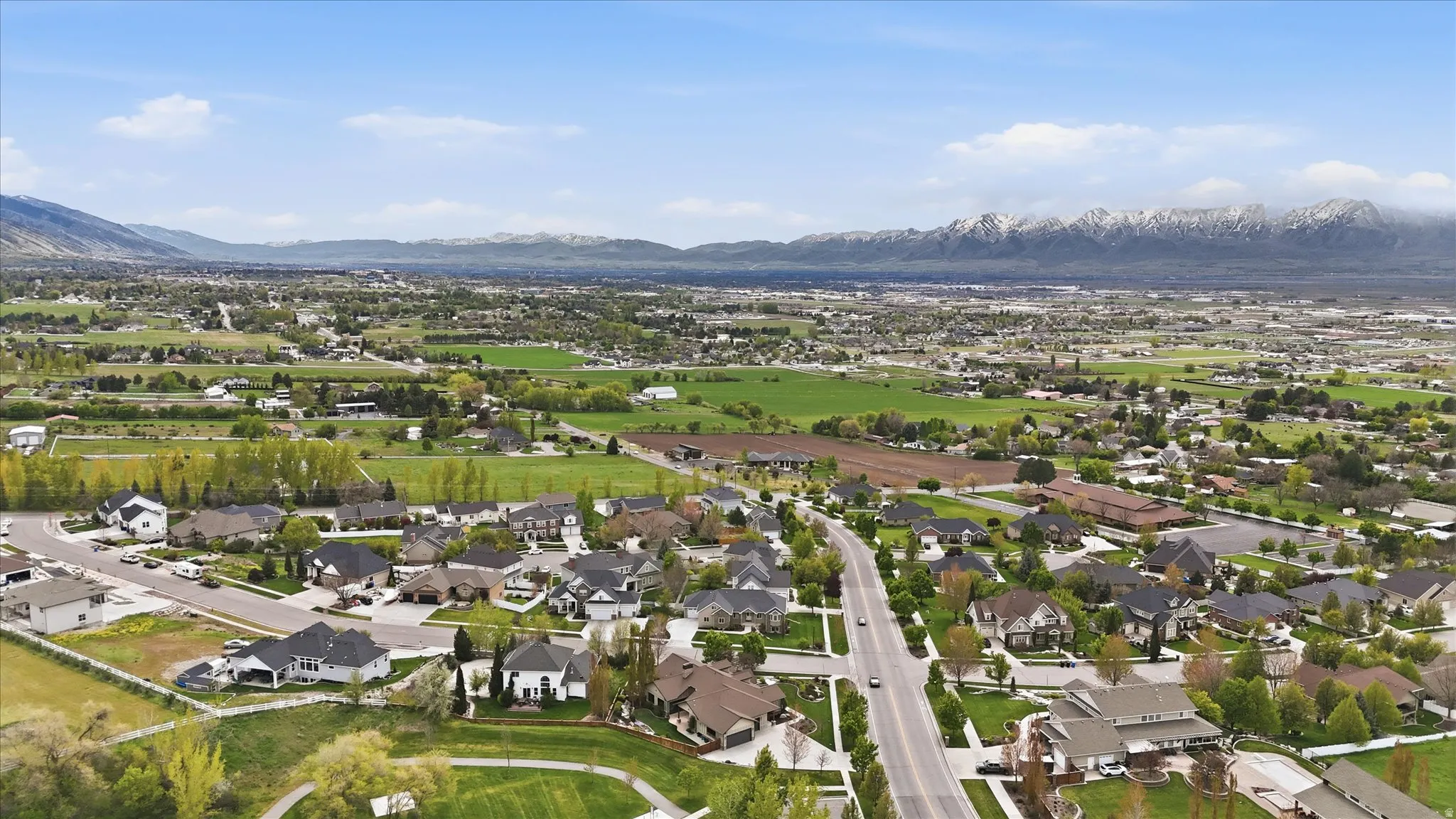 Expansive aerial perspective showcasing a residential development with varied architectural styles, surrounded by green fields and distant snow-capped mountains