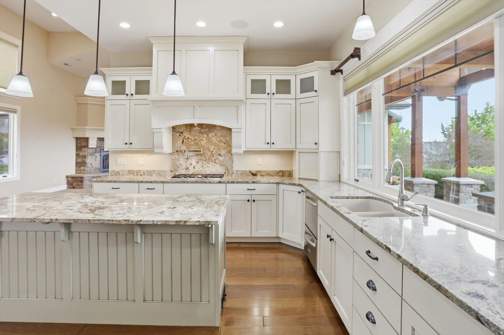 Expansive kitchen featuring a large central island with a beadboard base, light-toned granite countertops, white cabinetry with dark hardware, wood-finish flooring, and a large window overlooking an outdoor patio structure
