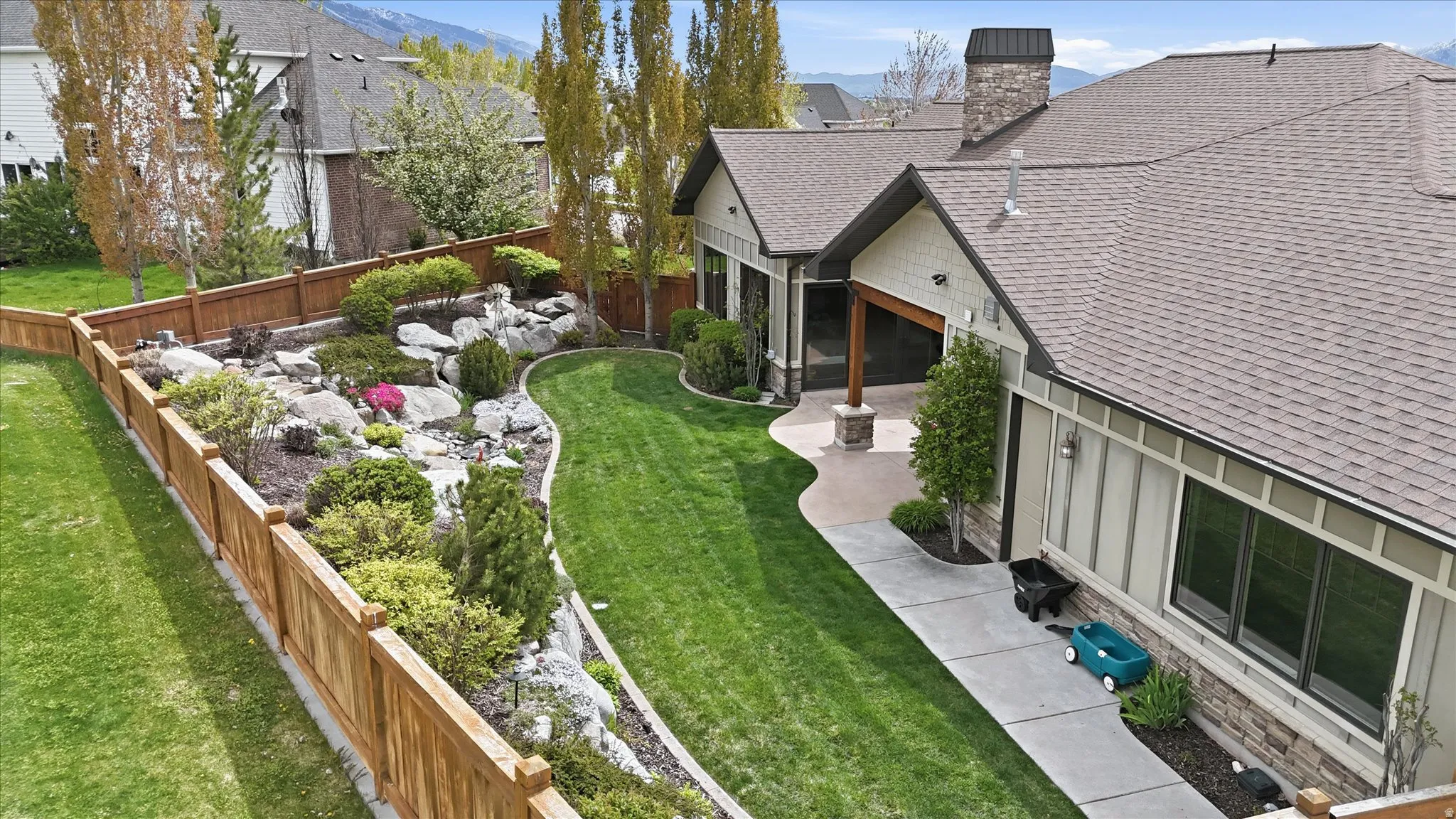 Expansive backyard featuring a manicured lawn, a rock garden with varied plantings, a wood privacy fence, a concrete patio, and a covered outdoor area with a stone-clad column