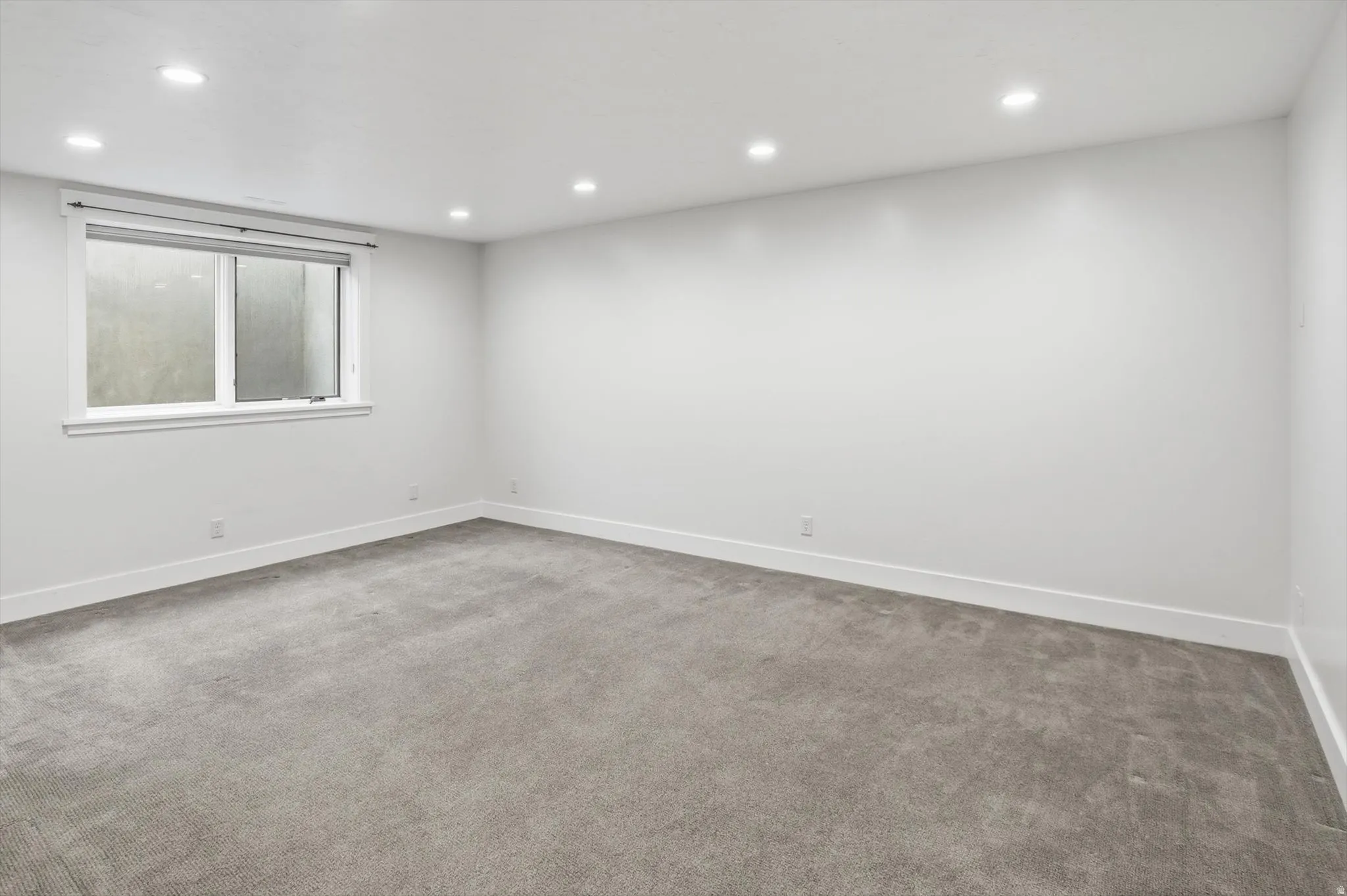 Spacious room featuring neutral carpeting, white walls, recessed ceiling lighting, a white window frame, and white baseboards