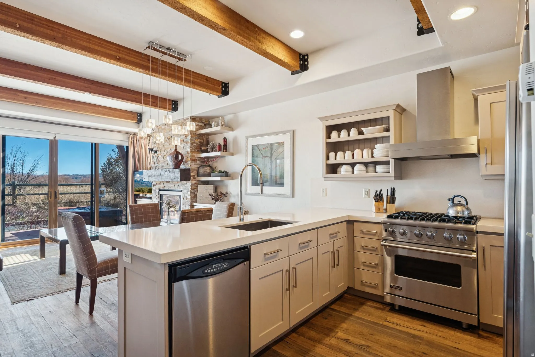 Kitchen featuring an expansive island with a stainless steel dishwasher, wood-finish flooring, exposed beam ceiling, stainless steel range with matching hood, and sliding glass doors opening to an outdoor patio