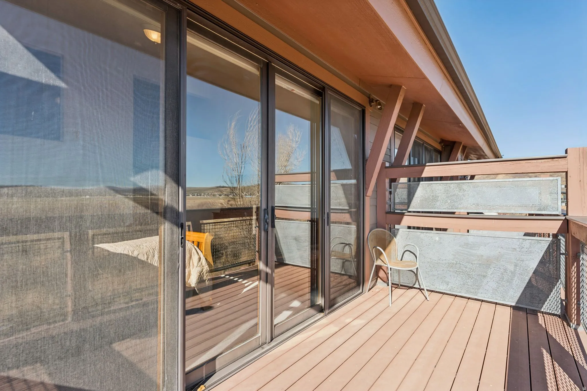 Exterior deck with wood-finish flooring, featuring sliding glass doors, a decorative metal railing, and an architectural overhang with exposed beams