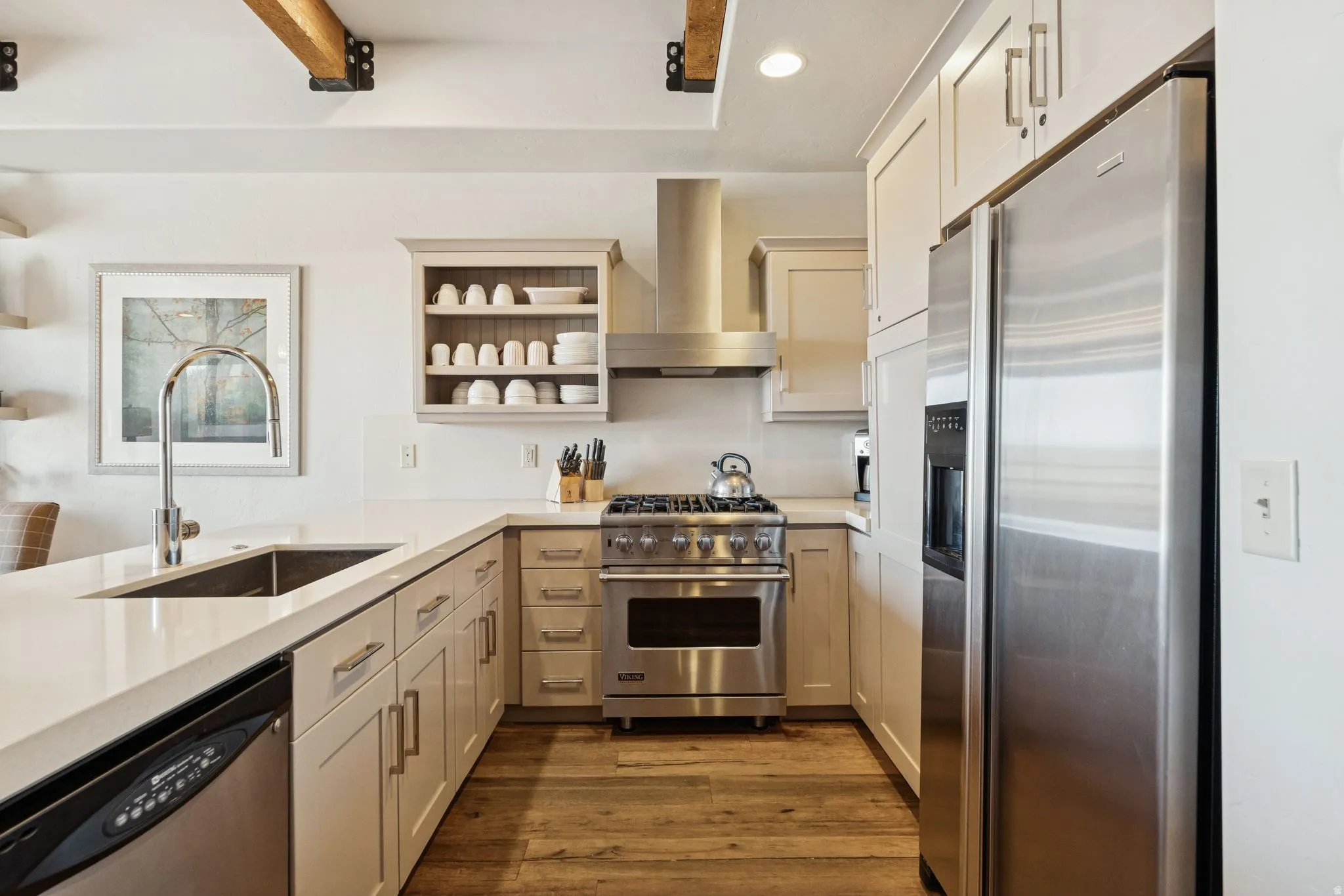Modern kitchen featuring exposed wood ceiling beams with black metal brackets, light-toned cabinetry, stainless steel appliances, a deep basin sink with a gooseneck faucet, and wood-finish flooring