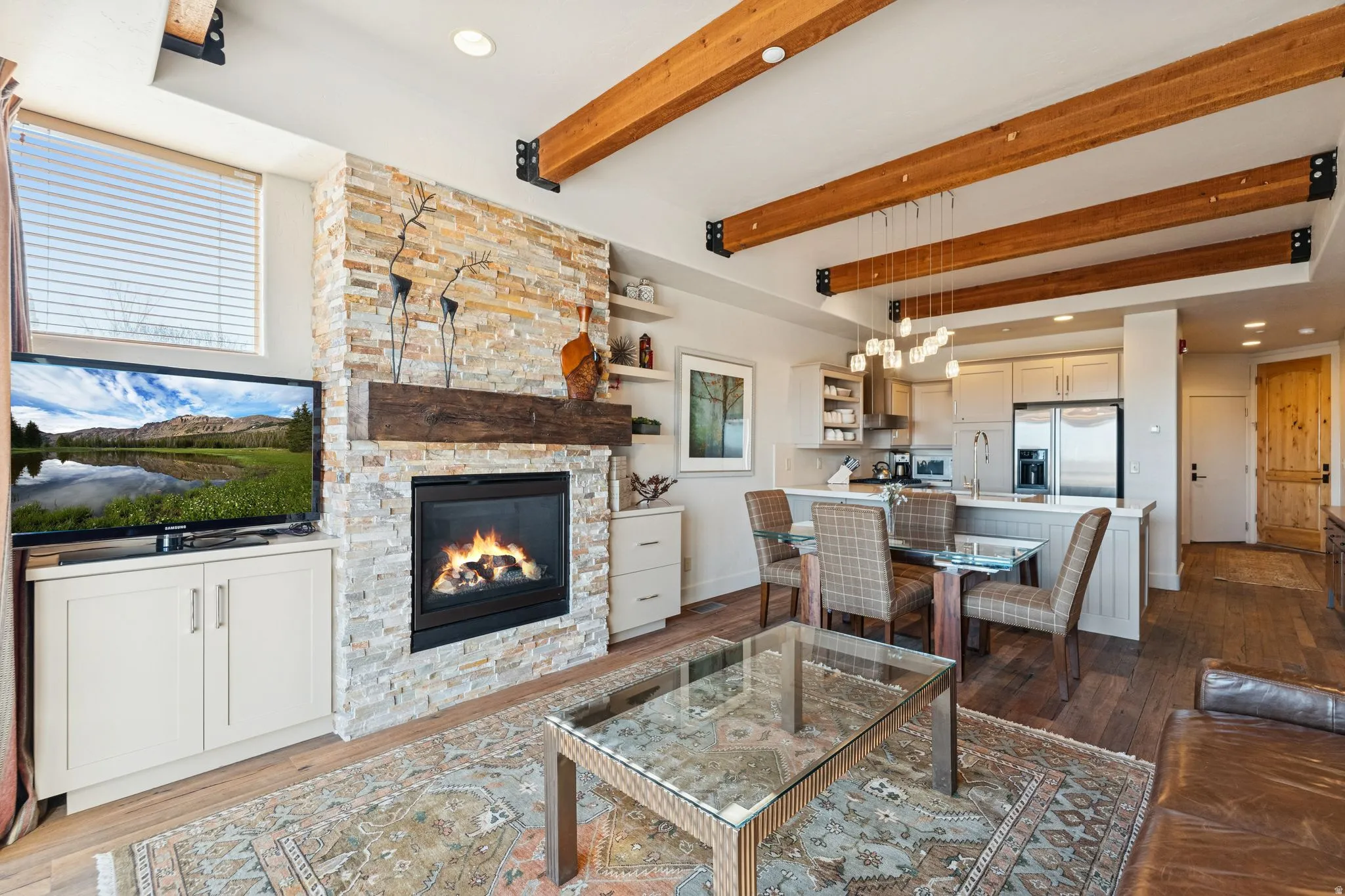 Open-concept living space featuring exposed wood beams, a stacked stone fireplace with a substantial wood mantle, and wood-finish flooring