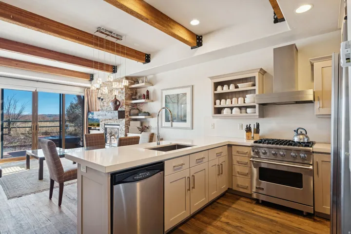 Kitchen featuring an expansive island with a stainless steel dishwasher, wood-finish flooring, exposed beam ceiling, stainless steel range with matching hood, and sliding glass doors opening to an outdoor patio