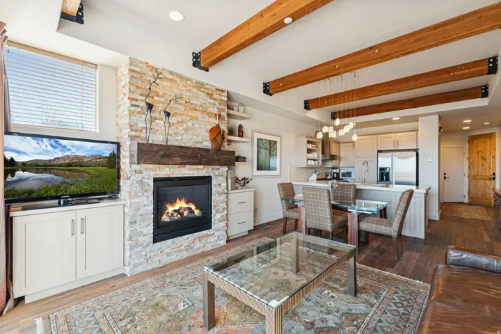 Open-concept living space featuring exposed wood beams, a stacked stone fireplace with a substantial wood mantle, and wood-finish flooring