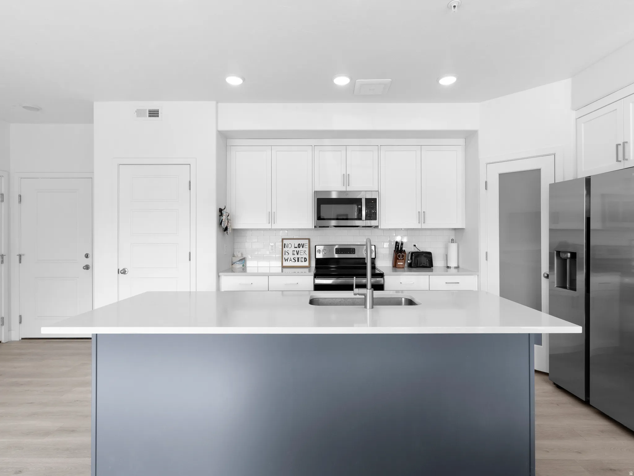 Kitchen featuring light wood-style flooring, stainless steel appliances, two tone cabinetry, an island with sink, and light stone countertops