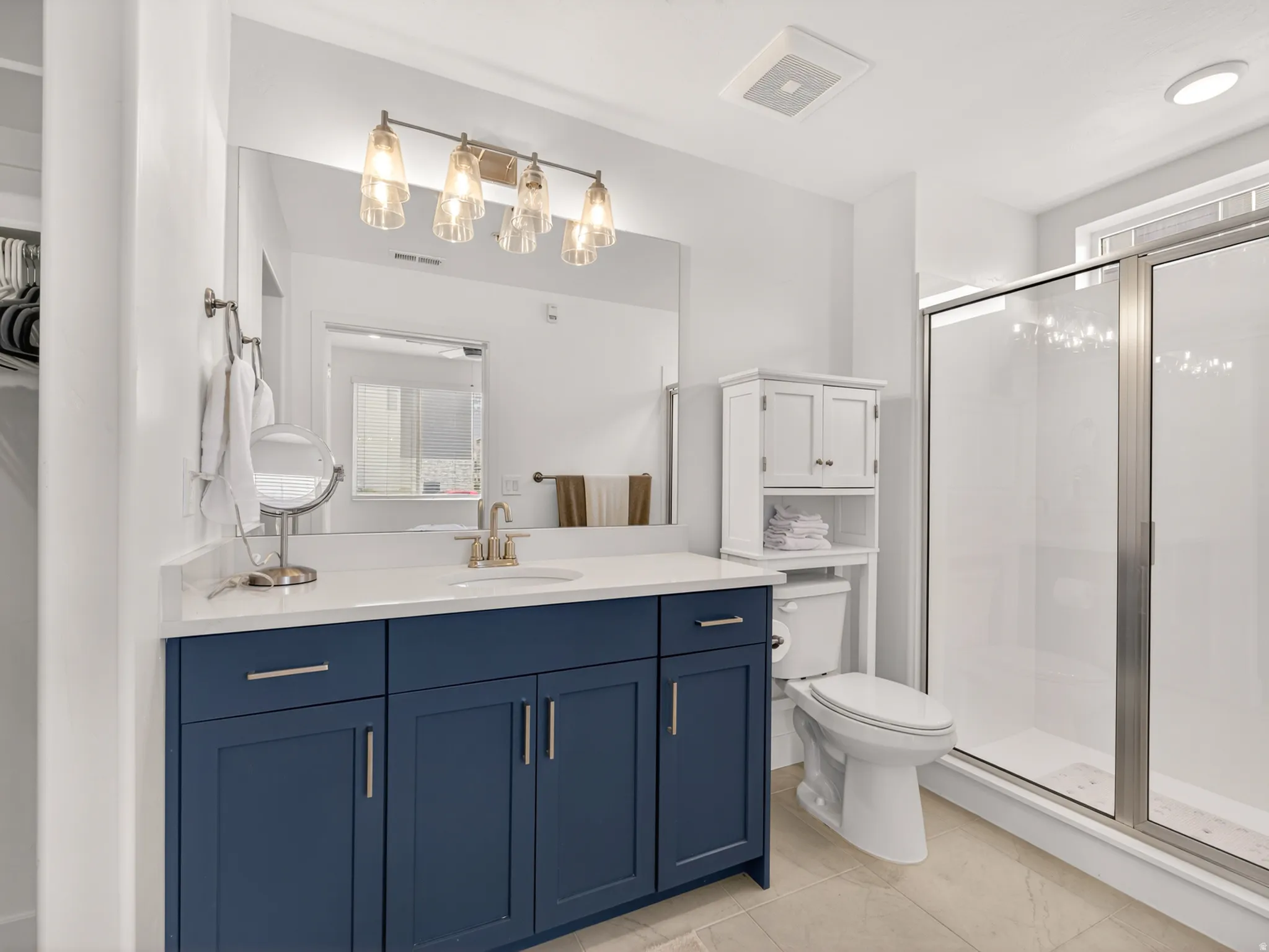 Bathroom featuring a navy blue vanity with a light-colored countertop, an overhead lighting fixture, a large mirror, a glass-enclosed shower, and light-toned floor tiling
