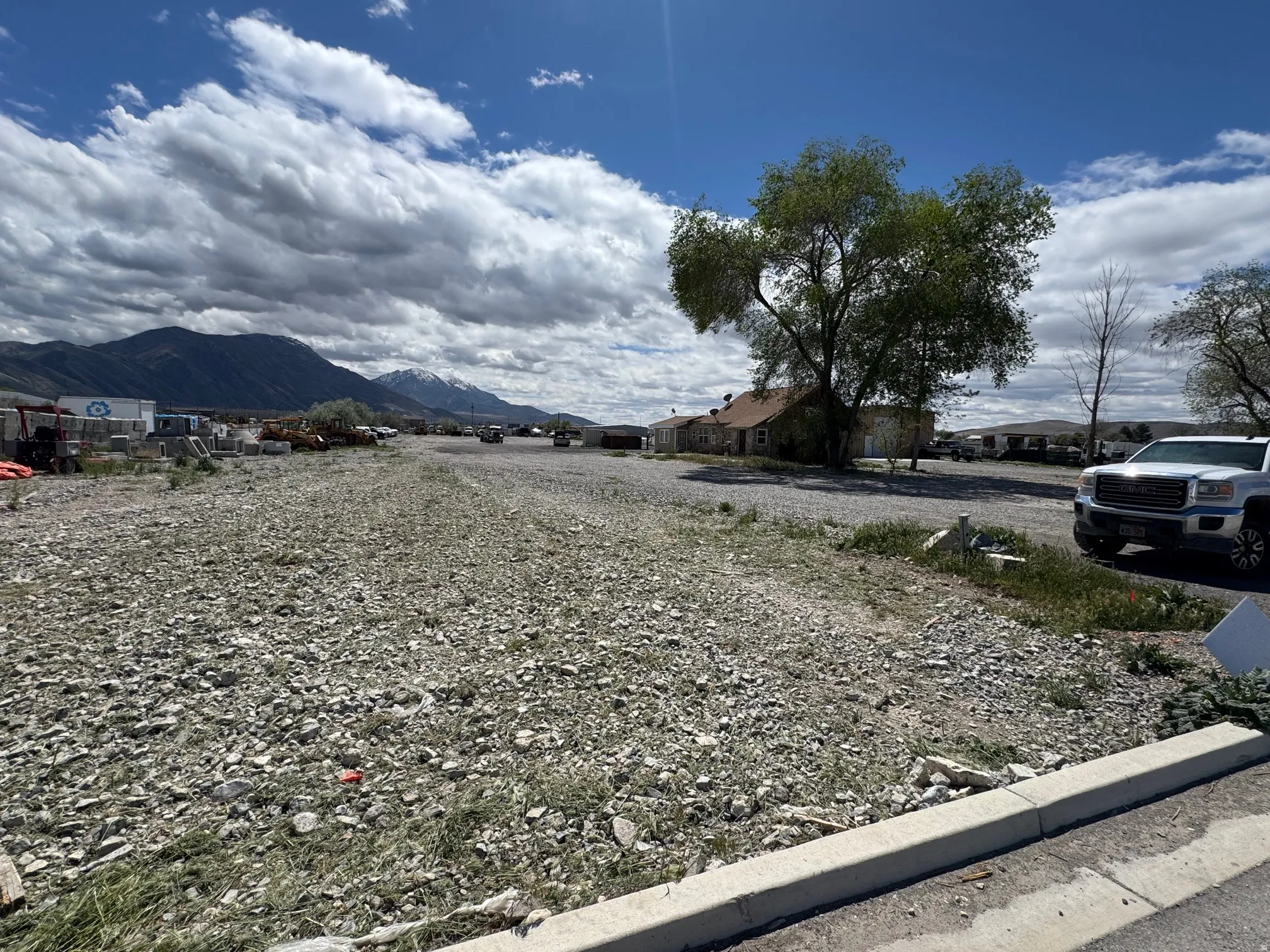 Expansive gravel lot featuring a paved curbline, mature tree, and distant mountain range
