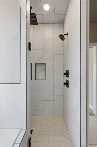Spacious walk-in shower featuring large format white tiled walls, a penny tile floor, a built-in shower niche, and matte black dual shower heads