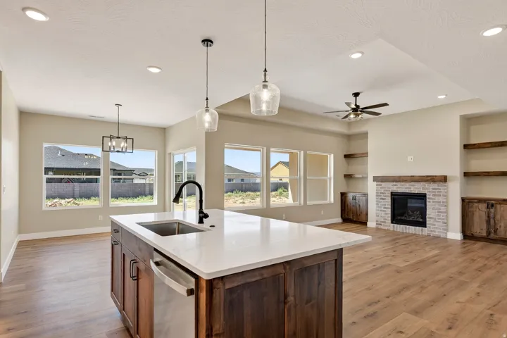 Open-concept living space with wood-finish flooring, a kitchen island featuring a stainless steel sink and dishwasher, a brick fireplace with a wood mantle, and built-in shelving with cabinetry