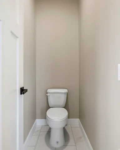 Contemporary water closet with a white toilet, light neutral wall color, white trim, and light-colored tiled flooring