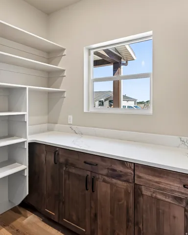 Spacious pantry featuring extensive built-in shelving, rich wood-finish base cabinetry, a crisp white countertop, and a window