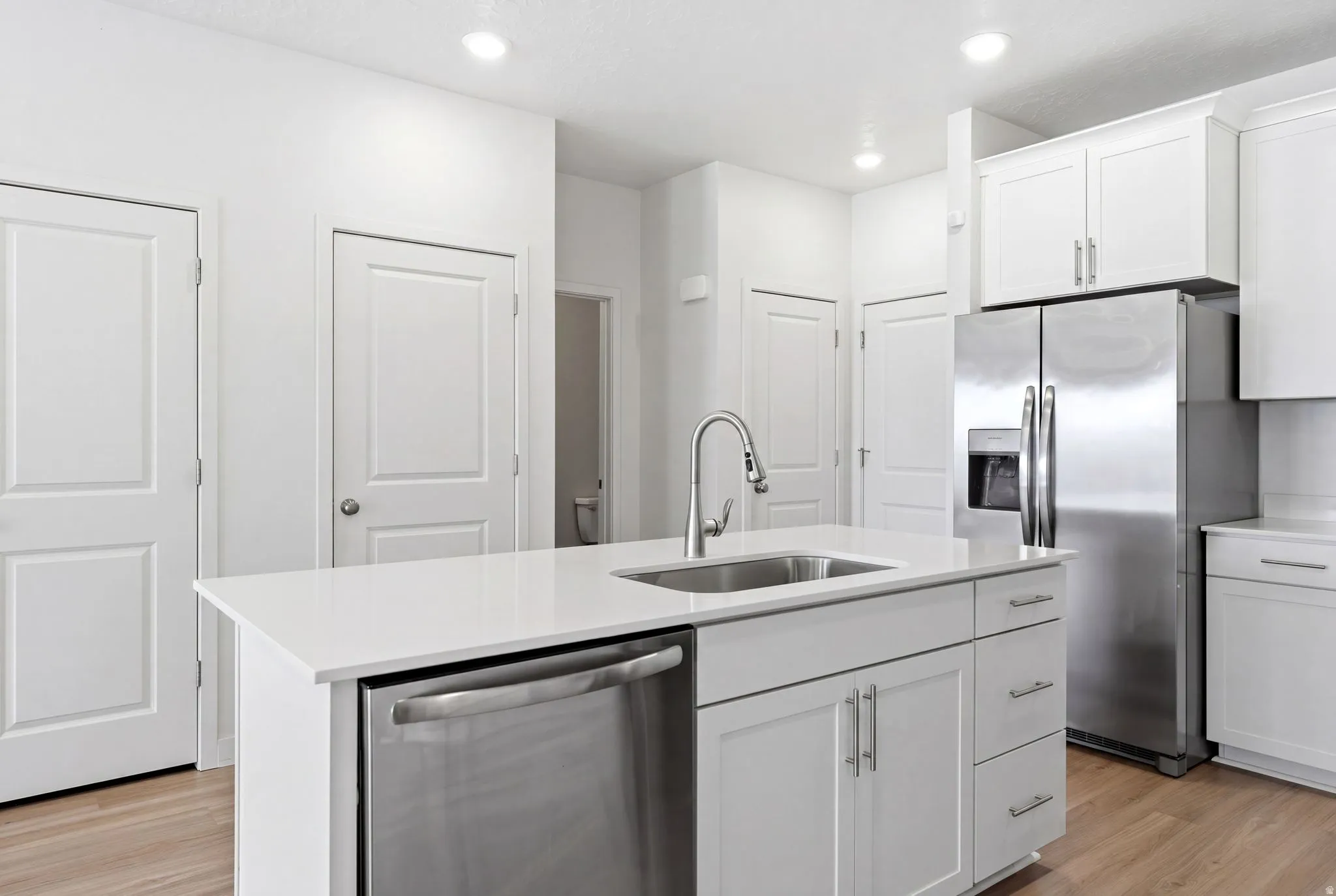 Kitchen featuring white shaker-style cabinetry, a central island with an undermount sink and stainless steel faucet, stainless steel appliances, and light wood-finish flooring