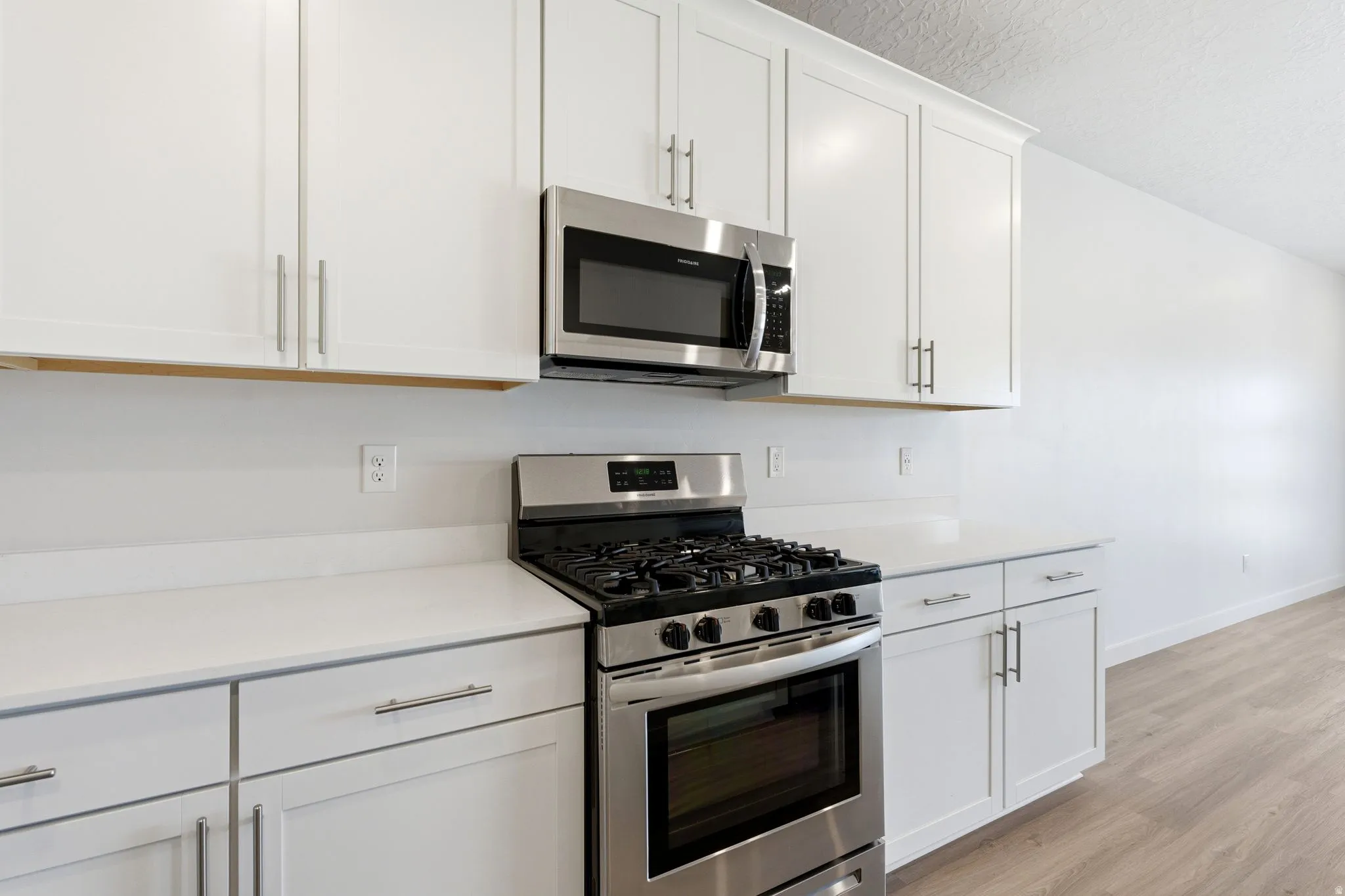 Kitchen featuring white cabinetry, stainless steel microwave, gas range, light-toned countertops, and wood-finish flooring