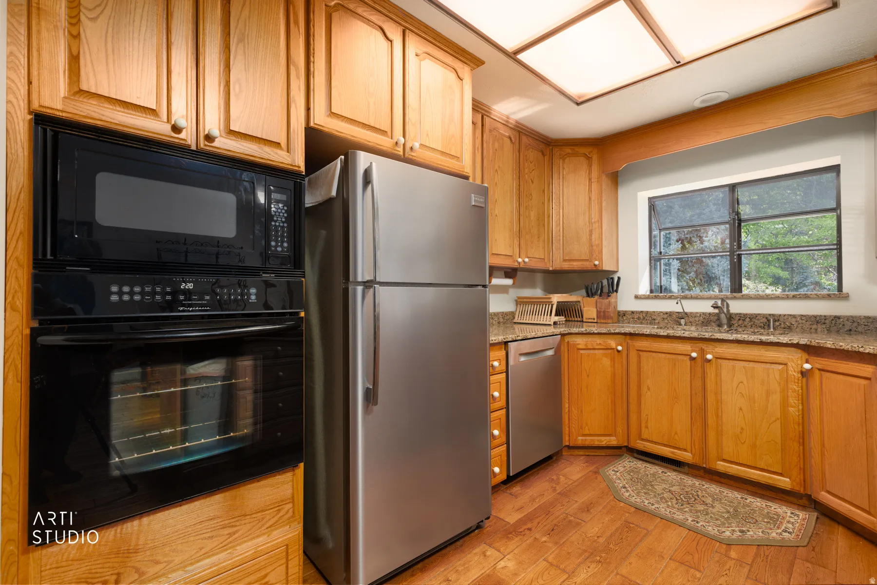 Kitchen featuring wood-finish flooring, light wood cabinetry, stainless steel refrigerator and dishwasher, black wall oven and microwave, and a window above the sink