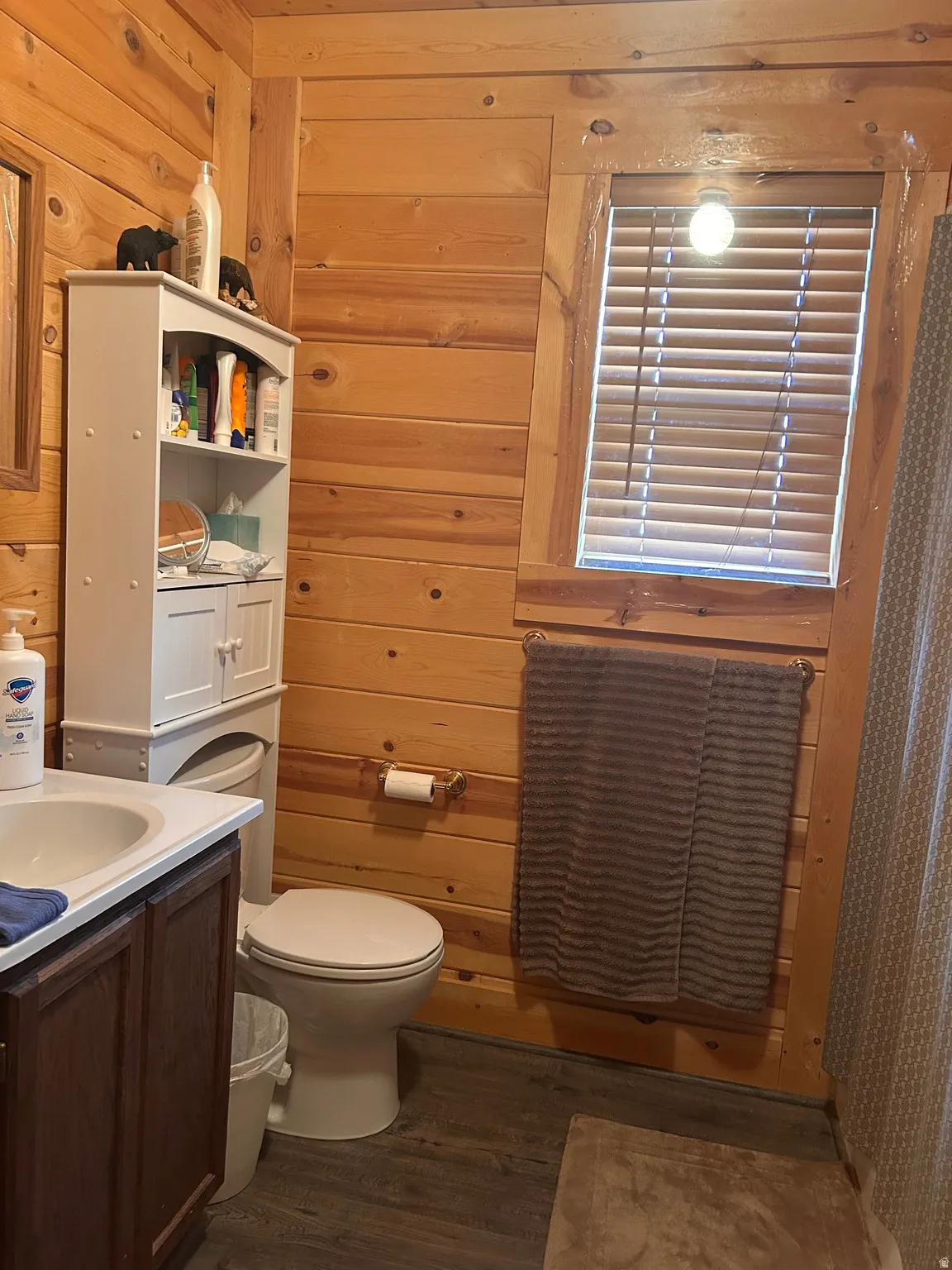 Bathroom featuring wood-finish paneling throughout, a vanity with a white basin and dark wood-finish cabinetry, a window with horizontal blinds, and wood-finish flooring