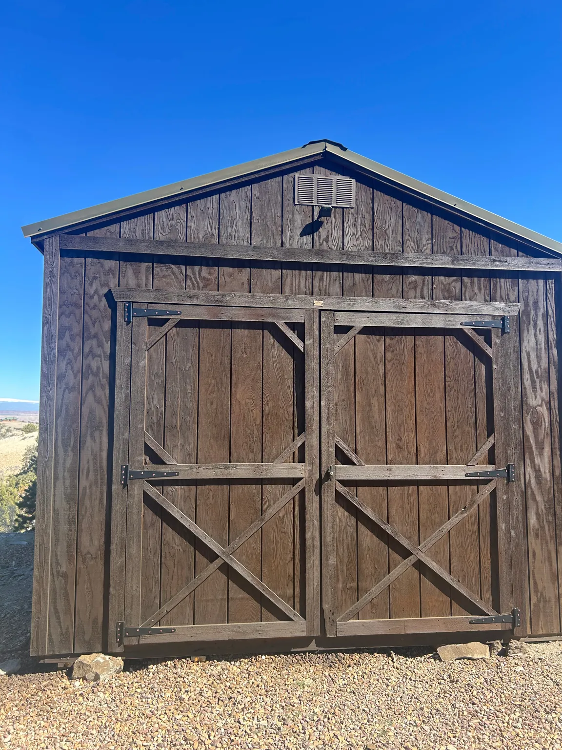 Gable-roofed shed with wood-finish siding, double barn doors featuring crossbuck detailing, and a louvered vent