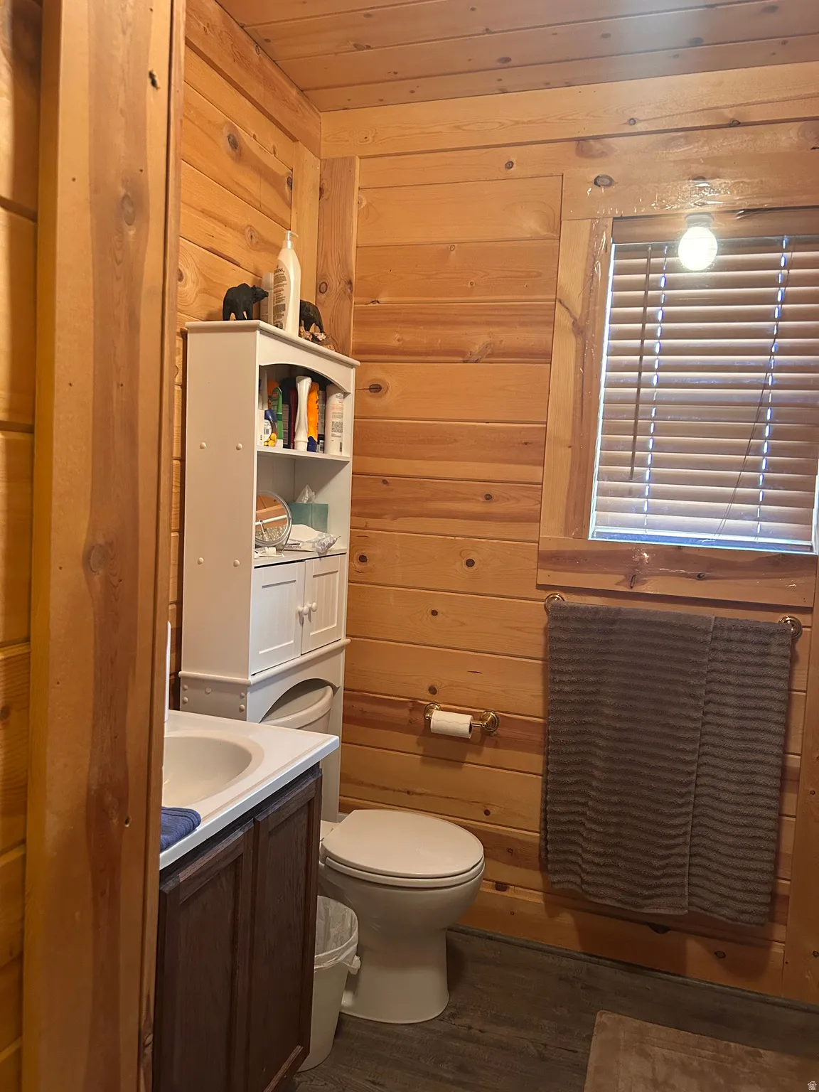 Bathroom featuring wood paneling throughout, a vanity with a white countertop and basin, a toilet, a window with blinds, and wood-finish flooring