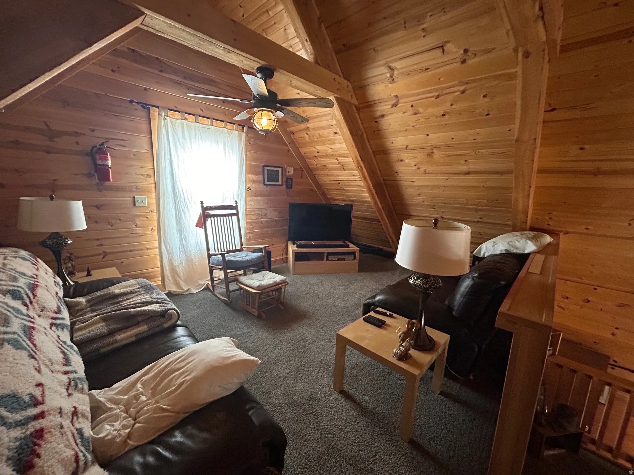 Attic space featuring wood plank walls and ceiling, exposed wood beams, and a ceiling fan with integrated lighting