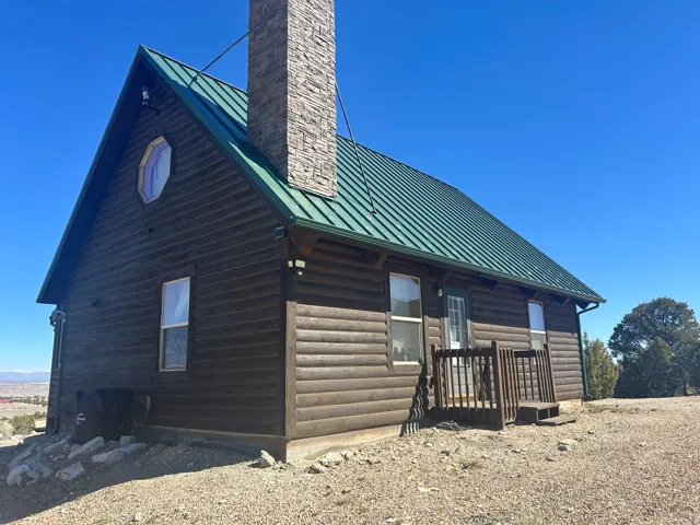 Log cabin exterior featuring a green metal roof, a stone-clad chimney, a small porch with wood railings, a unique octagonal window, and an exposed aggregate foundation