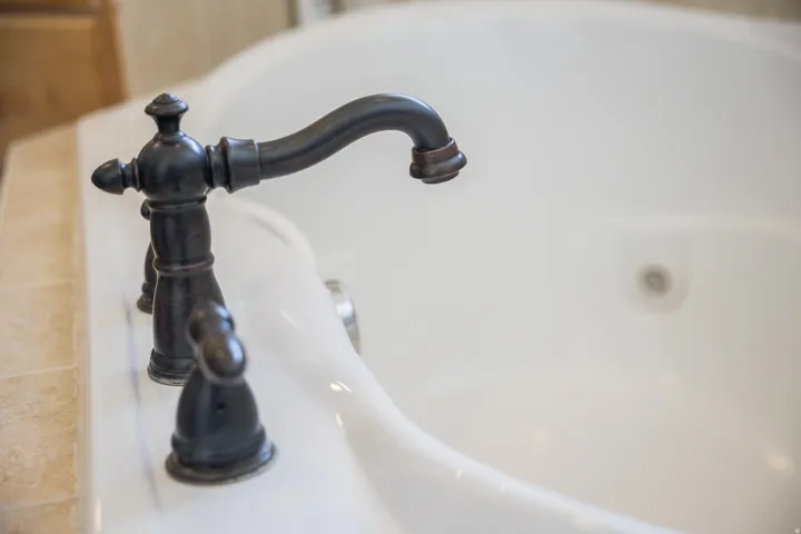 Oil-rubbed bronze tub filler with decorative handles and a curved spout, set into a tiled surround over a white soaking tub
