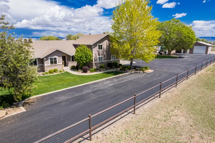 Expansive paved driveway leading to a tan stucco and stone facade home with a brown shingle roof, accompanied by a detached garage/shop building