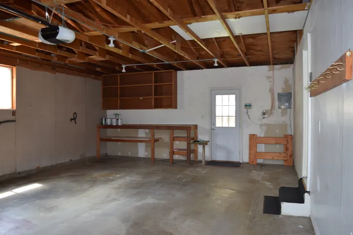 Spacious garage featuring exposed wood beam ceiling, concrete flooring, built-in shelving, work surface, and a single window