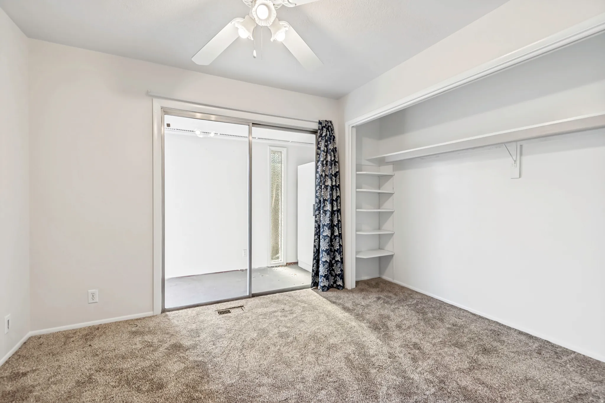 Carpeted interior space featuring a ceiling fan, sliding glass doors with an adjacent patterned curtain, and extensive built-in shelving