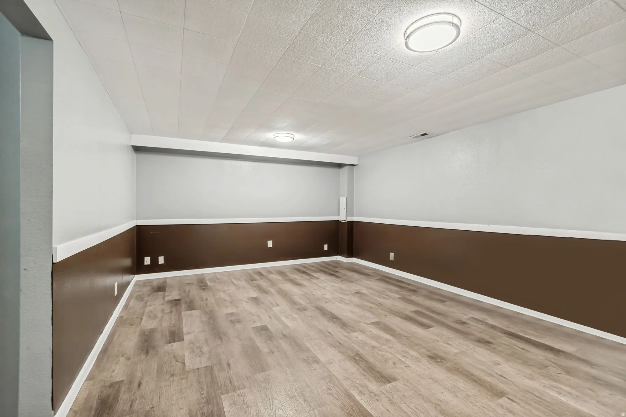 Spacious room featuring wood-finish flooring, two-tone wall paint with white trim, and a drop ceiling with integrated lighting
