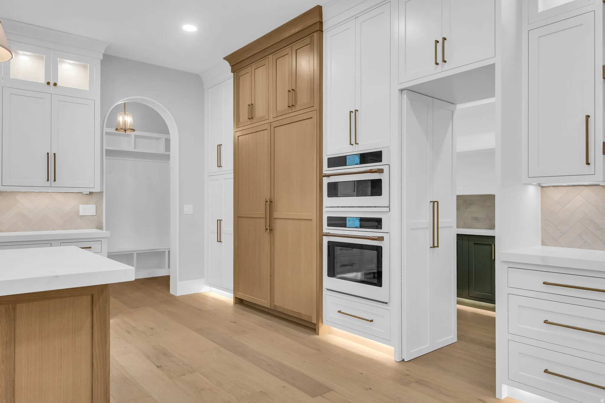 Gourmet kitchen featuring a built-in oven and microwave, custom cabinetry with brass hardware, wood-finish flooring, a herringbone pattern tile backsplash, and a white countertop