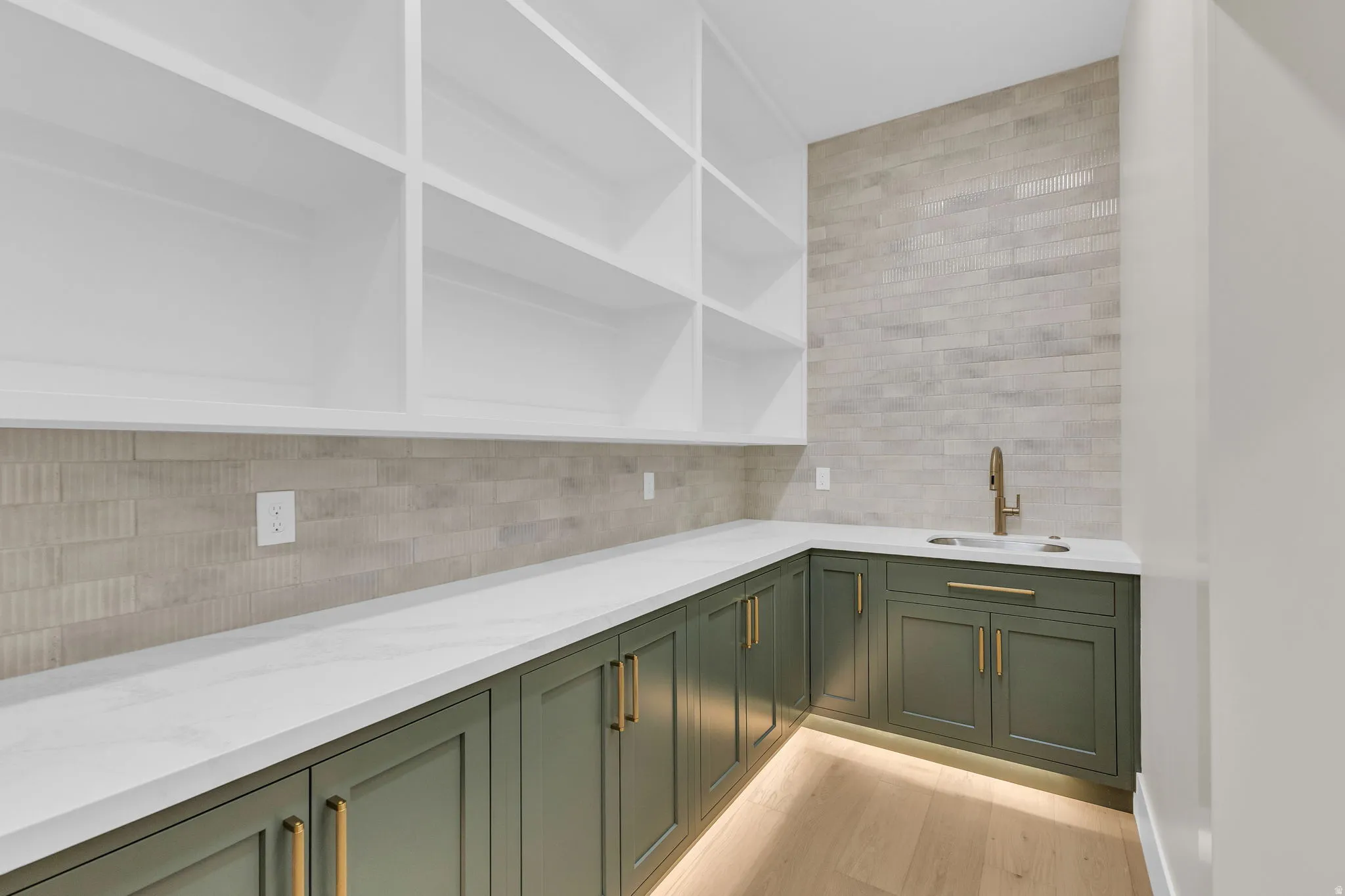Built-in cabinetry featuring white open shelving, a corner sink with a brass-finish faucet, dark green base cabinets with brass-finish hardware, and under-cabinet lighting, all complemented by light wood-finish flooring and a light-toned tiled backsplash
