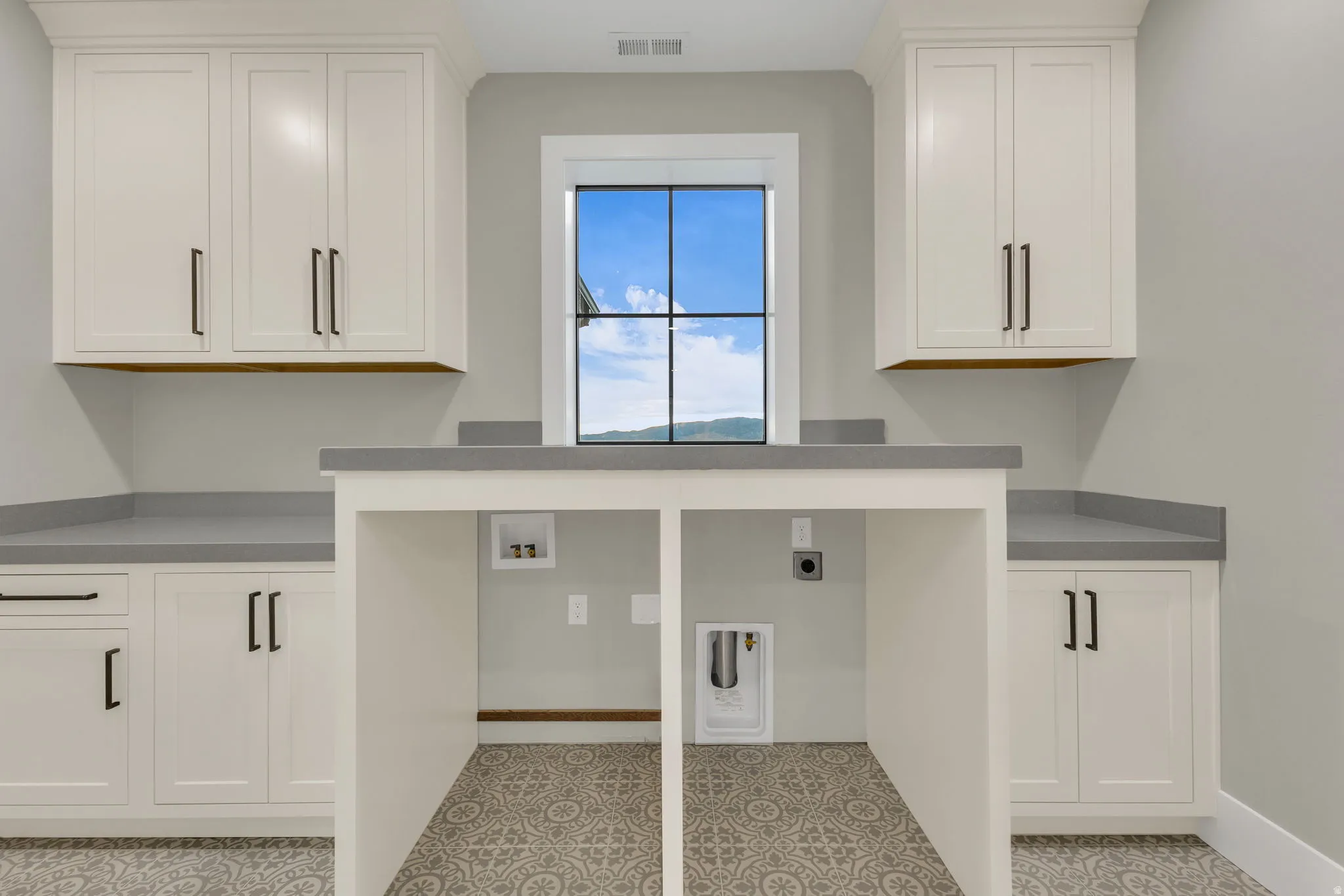 Laundry room featuring a large window, patterned floor tile, extensive cabinetry with dark hardware, and a built-in counter for appliances