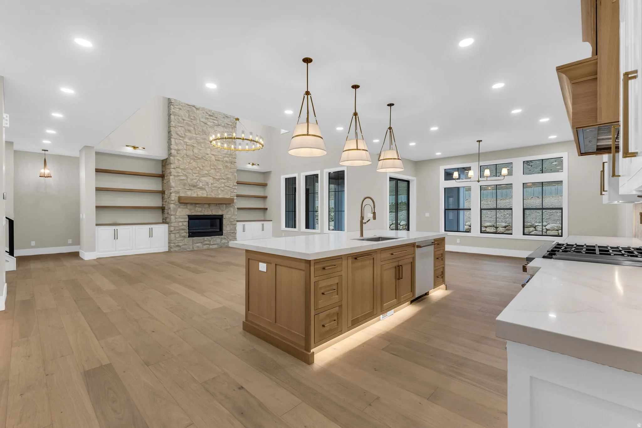 Open-concept living space featuring wide-plank wood-finish flooring, a stone-faced fireplace with a wood mantle, custom built-in shelving, a large kitchen island with a sink and pendant lighting, and multiple large windows