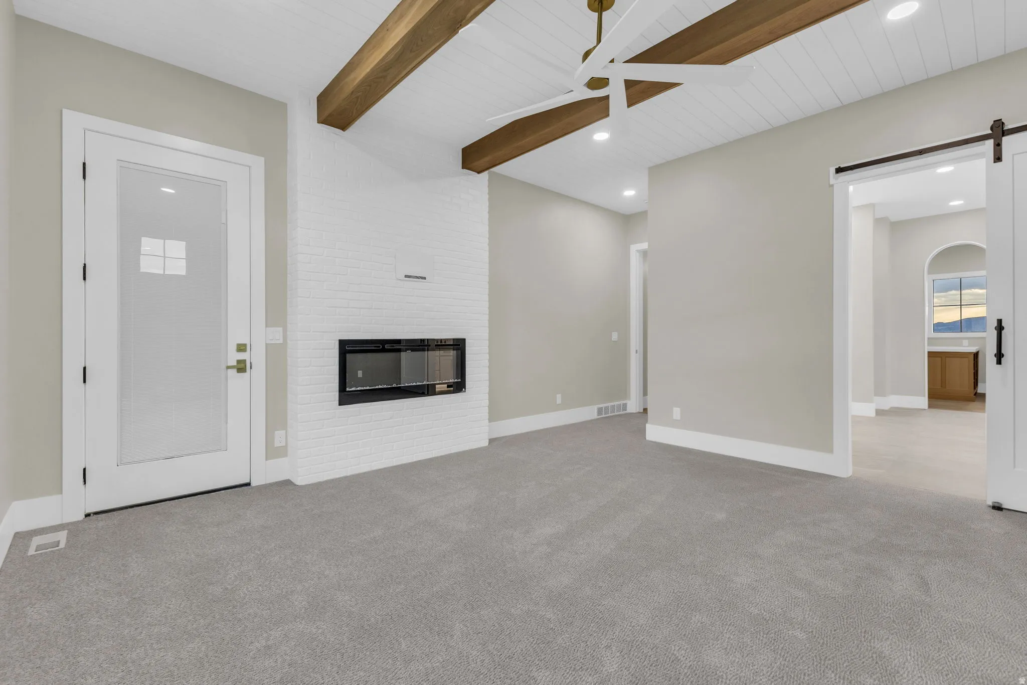 Spacious room featuring vaulted ceilings with exposed wood beams, a contemporary linear fireplace set into a white brick wall, and plush gray carpeting