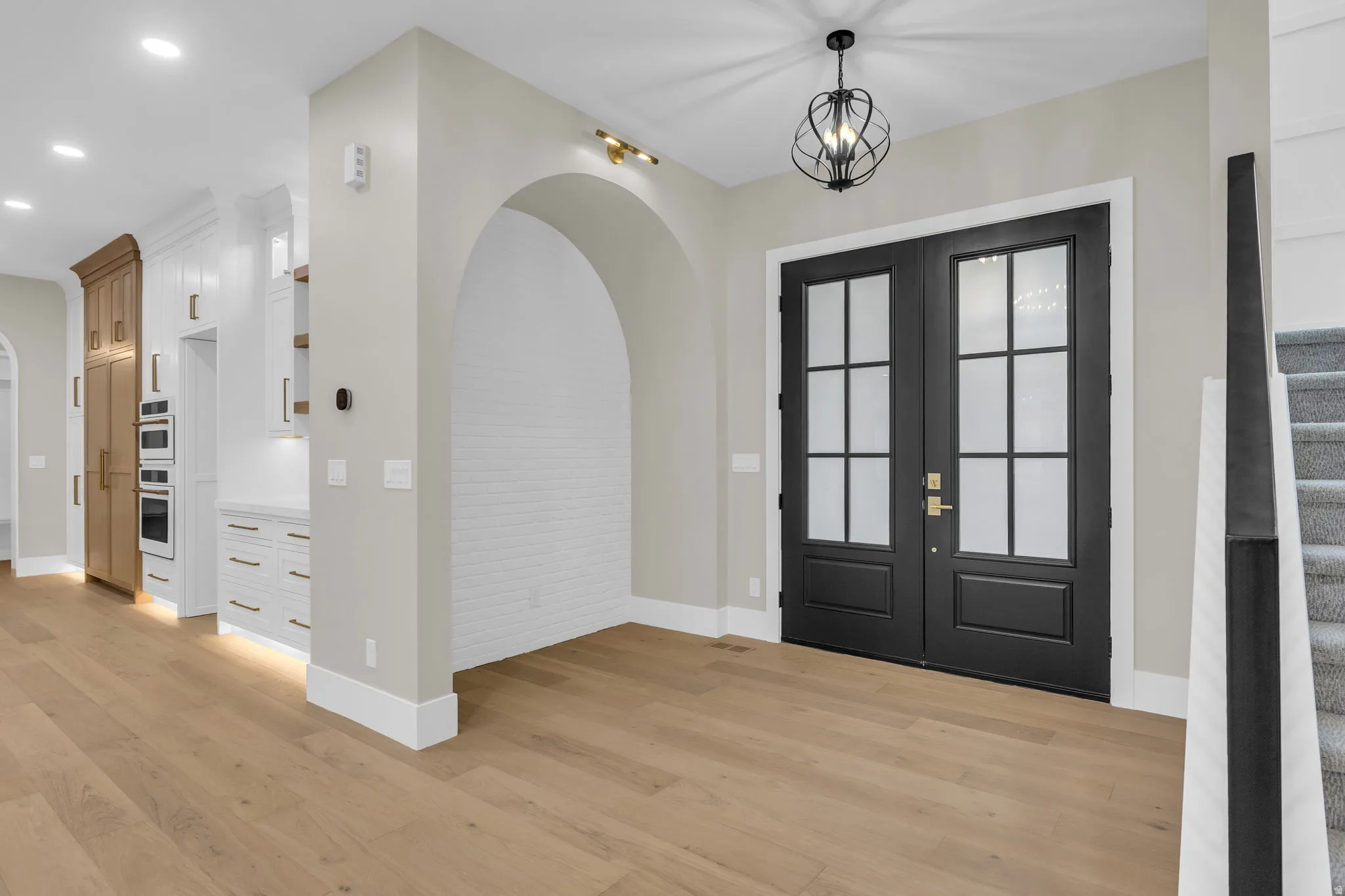 Double black entry doors with frosted glass panels, an arched alcove with white brick-patterned wall, light wood-finish flooring, and a contemporary chandelier