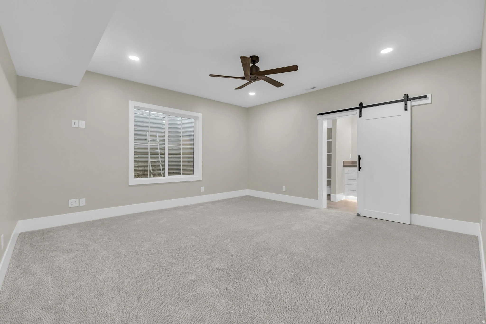 Spacious room featuring light gray carpeting, recessed lighting, a modern ceiling fan, a white barn door, and a horizontal window with white trim