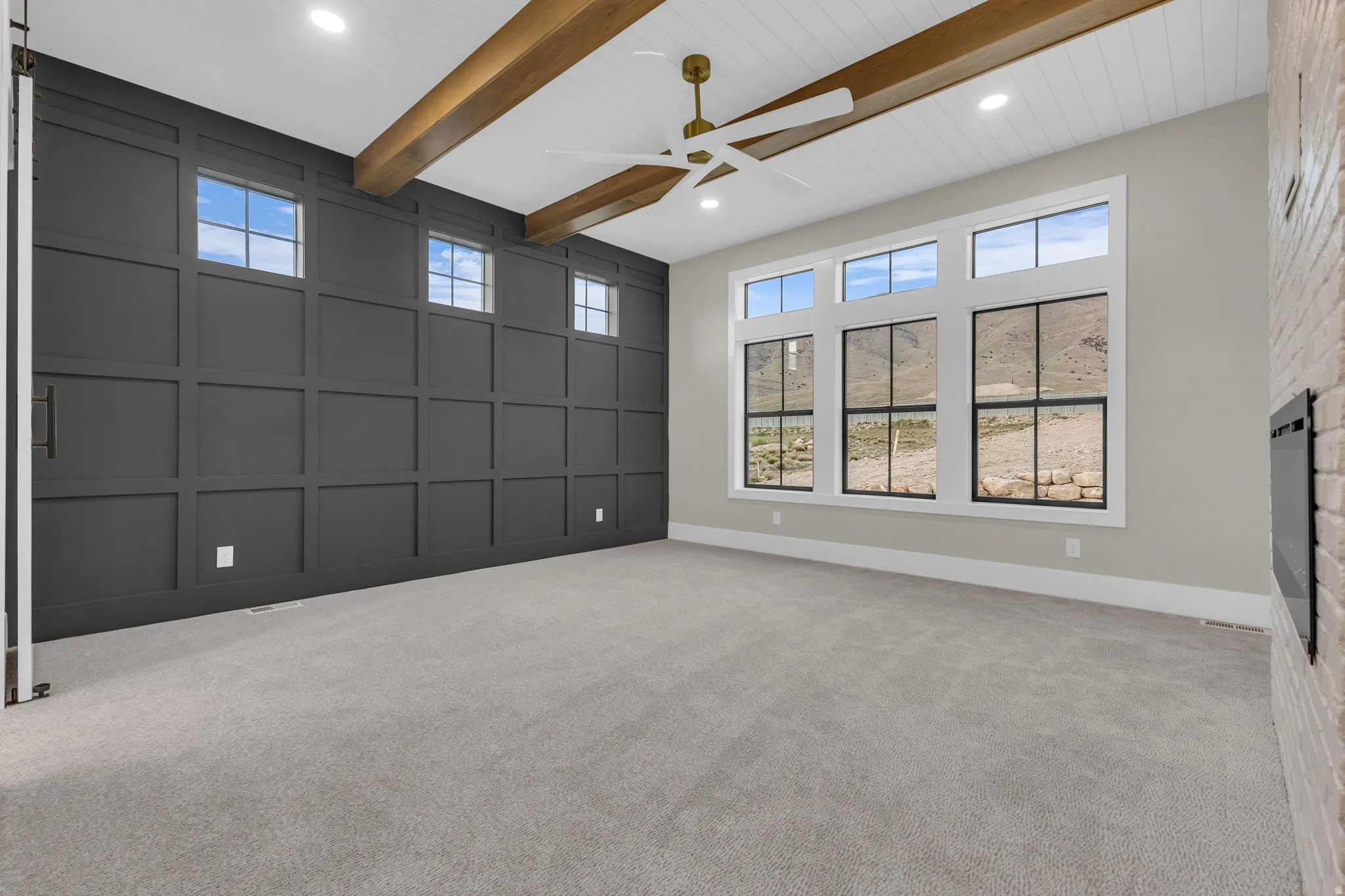 Spacious interior featuring a large window array with black frames, wood-beamed ceiling, recessed lighting, and a white brick fireplace
