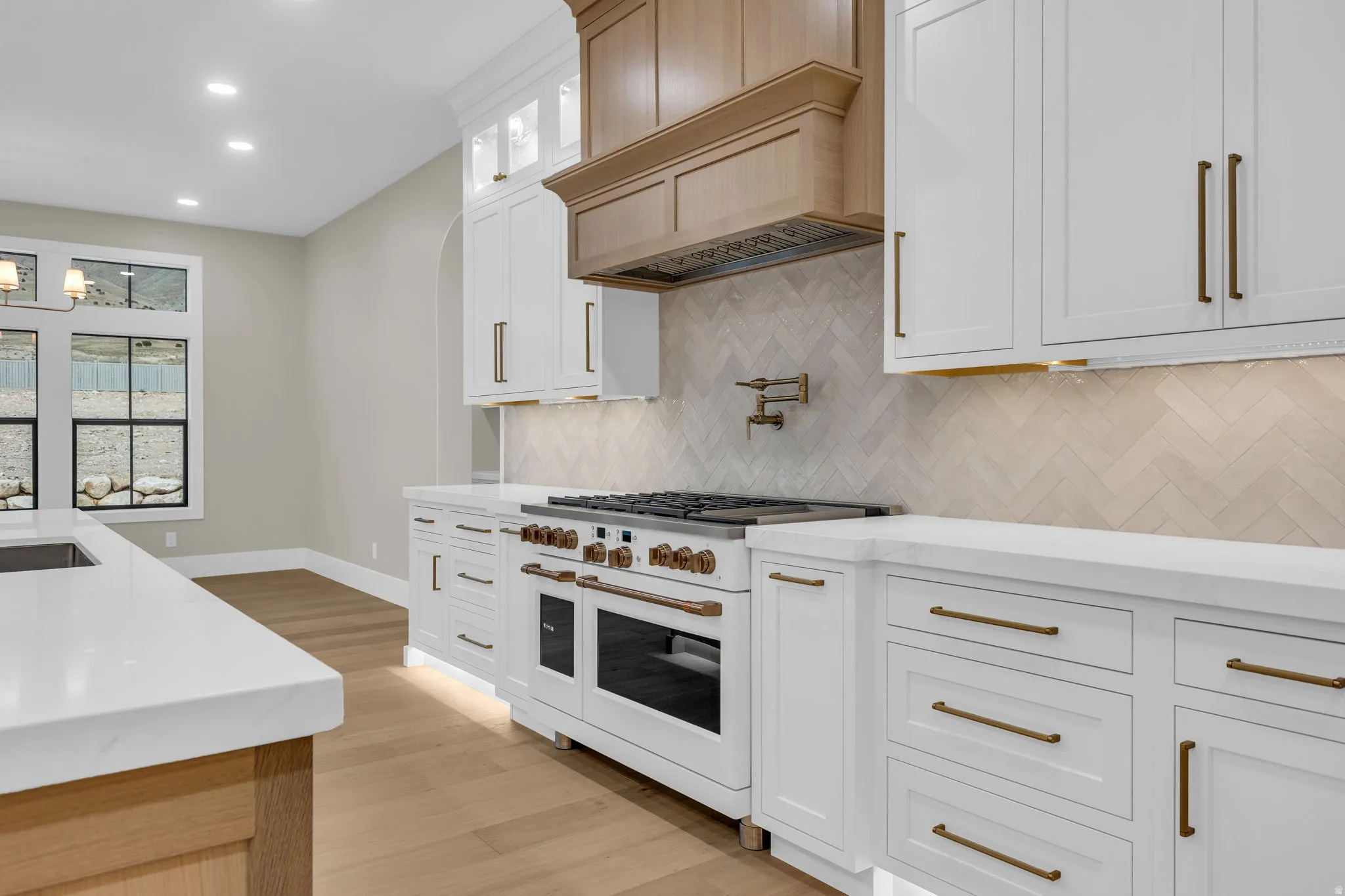 Gourmet kitchen featuring a large professional-grade range, white perimeter cabinetry with brass-toned hardware, light wood-finish flooring, and a herringbone pattern tile backsplash