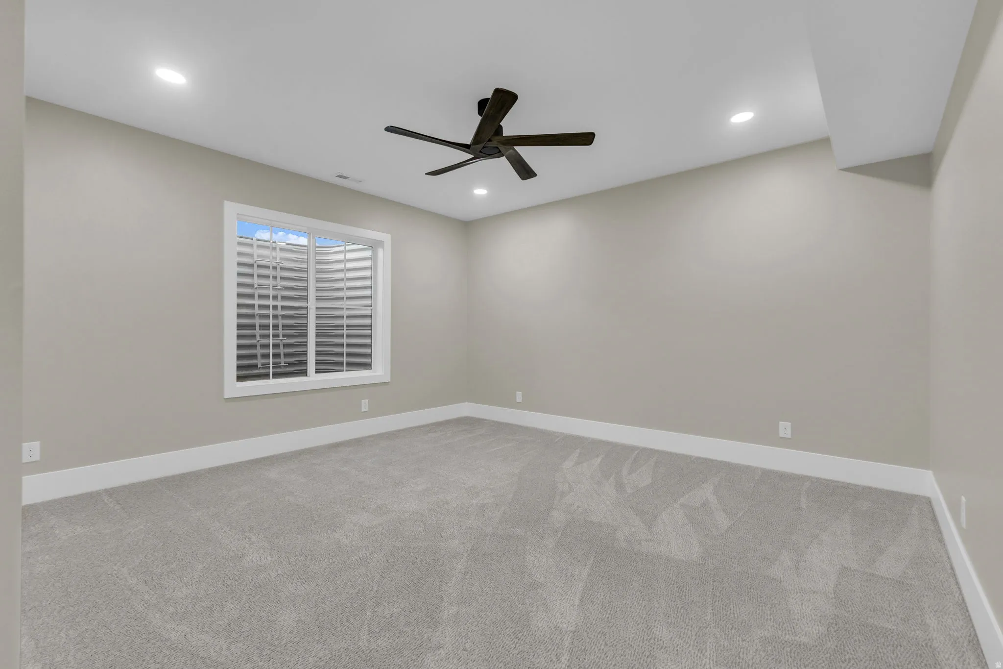 Carpeted room featuring a ceiling fan, recessed lighting, and a white-framed window with a window well
