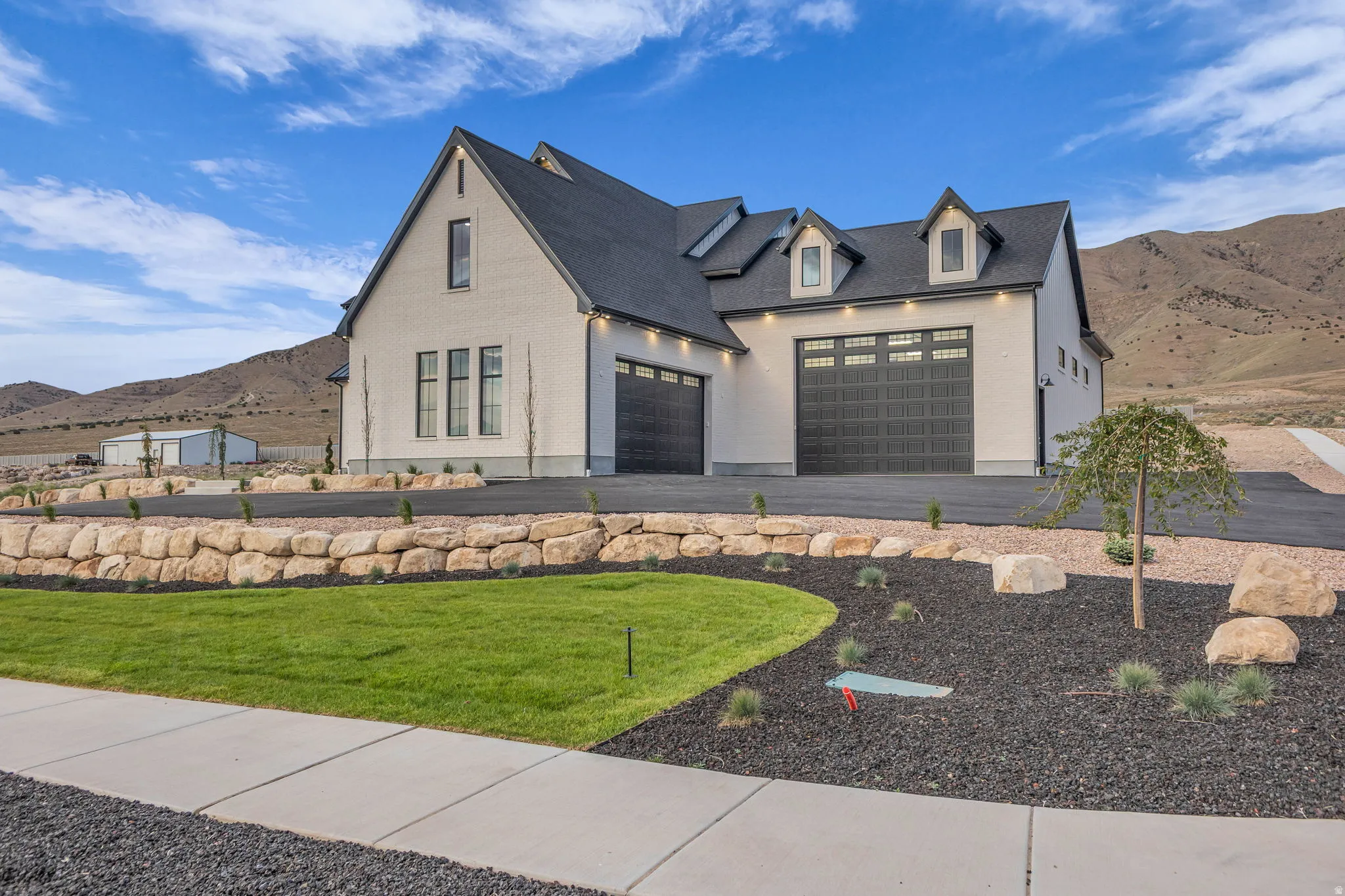 White facade architecture featuring multiple dormer windows, a dark shingle roof, and dark multi-pane garage doors