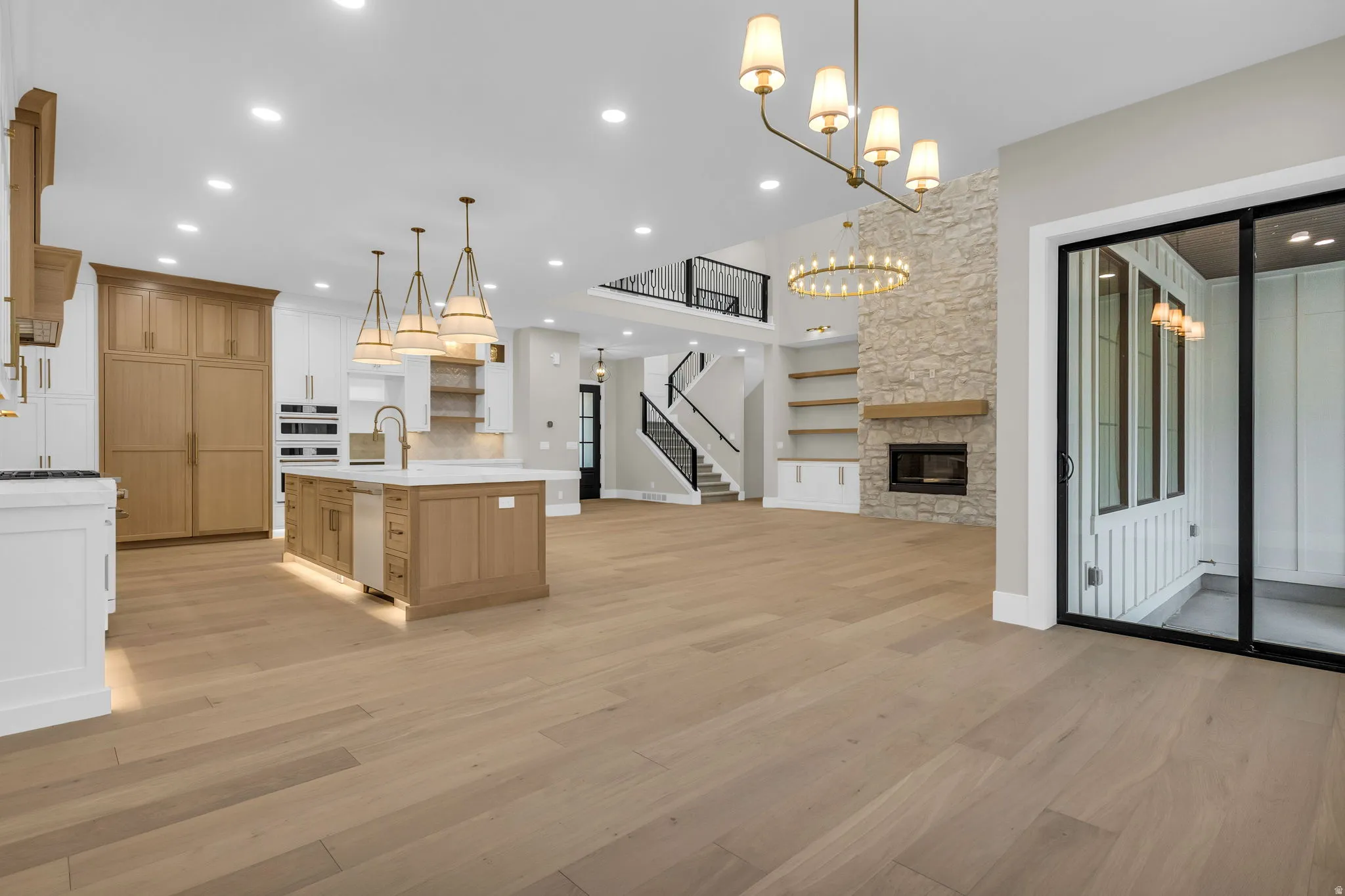 Open-concept living space with light wood-finish flooring, a stone-clad fireplace, a kitchen island with integrated sink, a two-story ceiling, and a sliding glass door