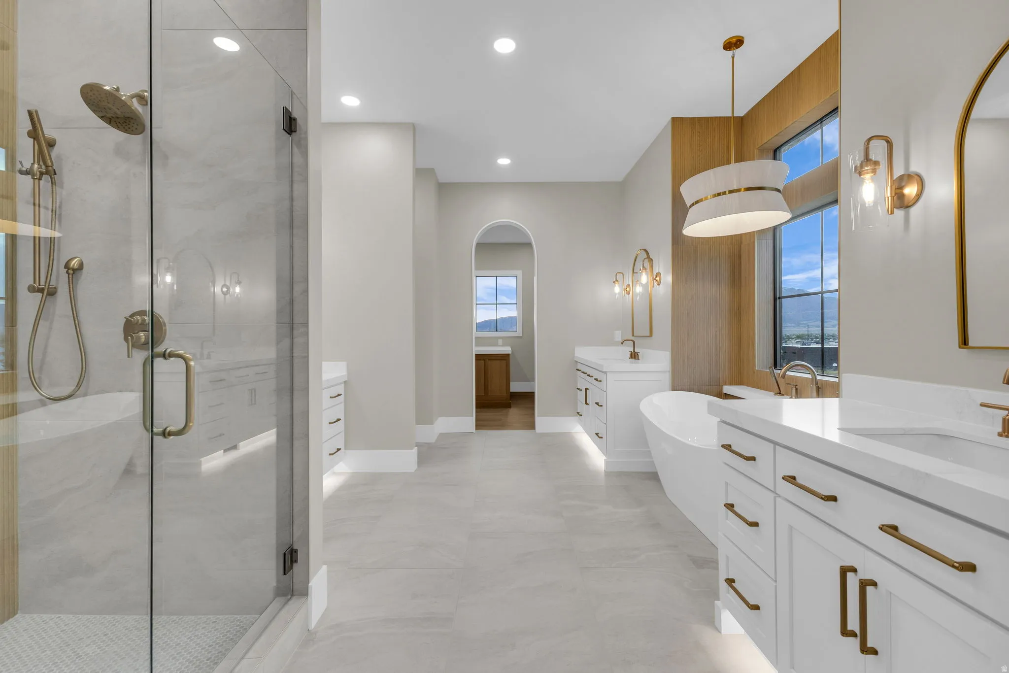 Sleek bathroom featuring a glass-enclosed shower with brushed brass fixtures, a freestanding soaking tub, dual vanities with white countertops and cabinetry, and large windows framed by wood-finish accents